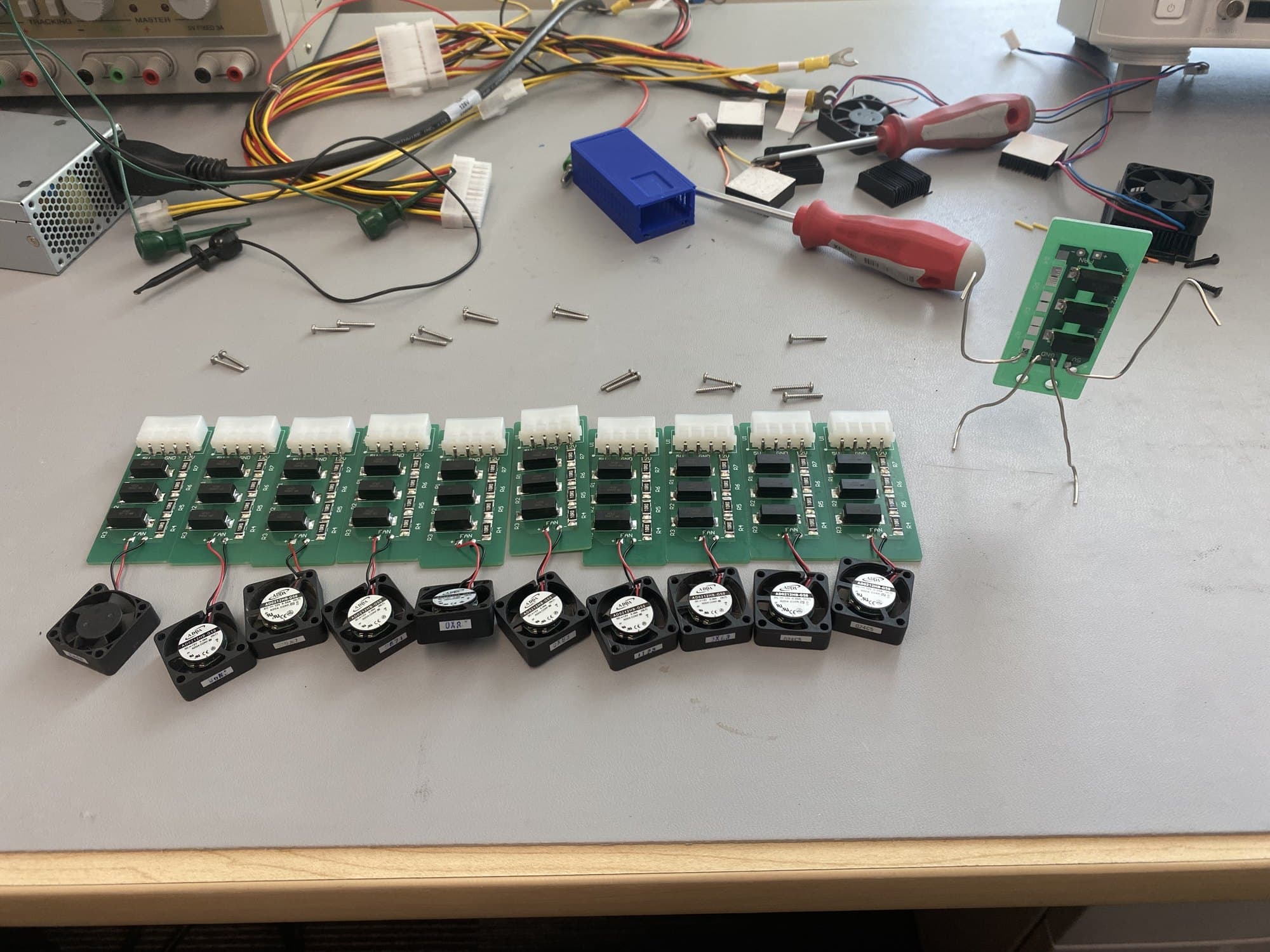 A row of finished green load boards on a lab bench, each with a small black fan wired to its fan pads, alongside an ATX supply, wiring harnesses and a blue 3D-printed enclosure